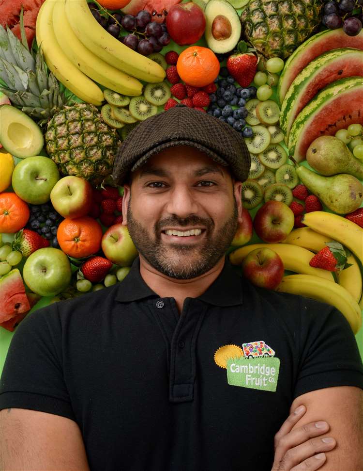 A head and shoulders image of Neil Bharadwa who is smiling to camera, wearing a Cambridge Fruit Company polo shirt and a flat cap. The background is a vast array of fruits.
